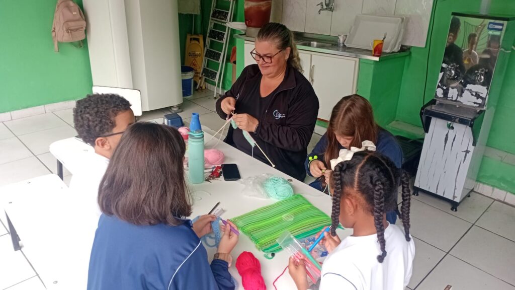 Grupo de alunos do 3º ano reunidos ao redor da mesa com novelos de lã, agulhas de crochê e trabalhos em andamento